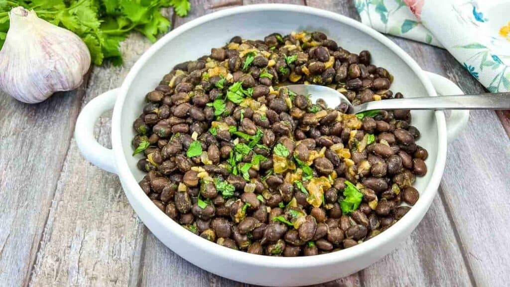 A white bowl of cooked black beans garnished with chopped herbs, placed on a wooden surface with a spoon in it. Garlic and cilantro are visible in the background.