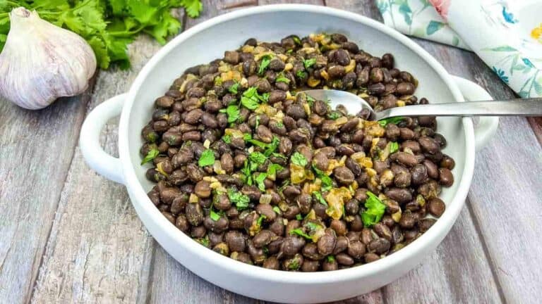 A white bowl of cooked black beans garnished with chopped herbs, placed on a wooden surface with a spoon in it. Garlic and cilantro are visible in the background.