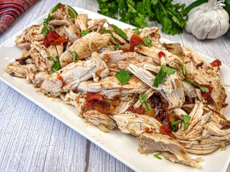 A plate of shredded, cooked chicken garnished with chopped herbs and pieces of tomato on a wooden surface. Garlic and cilantro in the background.