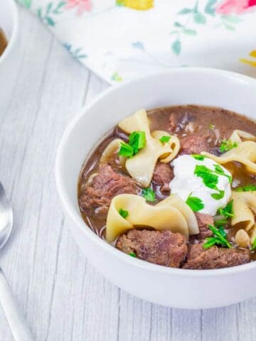 Bowl of beef stroganoff soup with sour cream and parsley garnish on a light wooden table next to two spoons and a floral napkin.