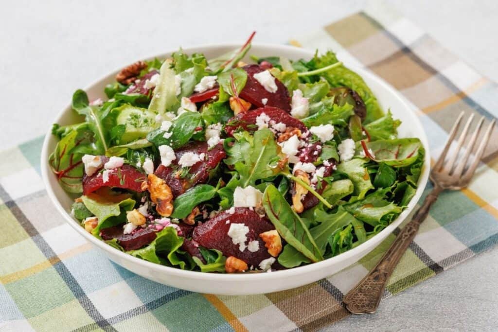 A fresh salad with mixed greens, sliced beets, walnuts, and crumbled feta cheese in a white bowl, placed on a checkered tablecloth with a metal fork beside it.