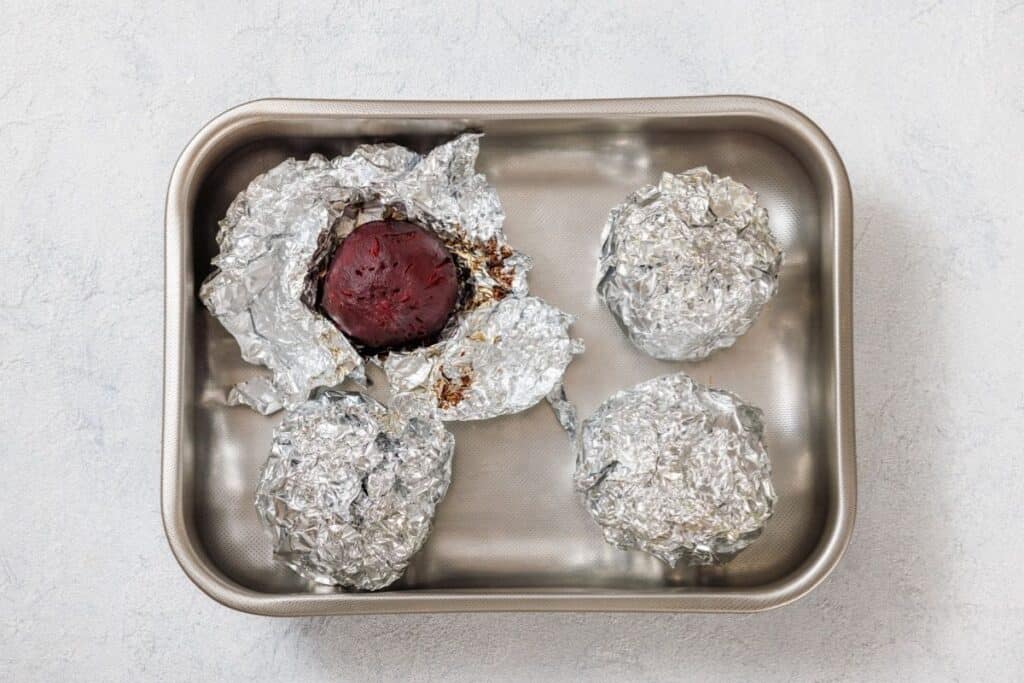 Four foil-wrapped items in a baking pan, with one partially unwrapped to reveal a red beet.