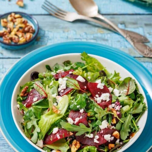 A bowl of salad with mixed greens, sliced beets, walnuts, and crumbled cheese on a blue rimmed plate. Silver cutlery and a small bowl of walnuts are in the background.