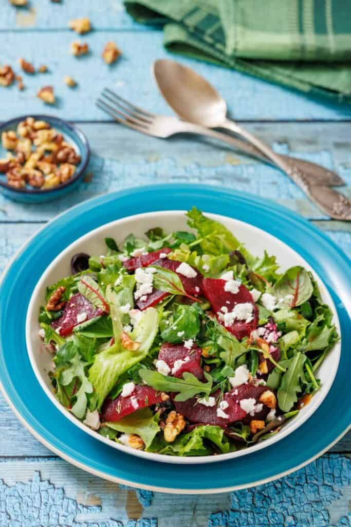 A salad with mixed greens, roasted beets, feta cheese, and walnuts in a blue-rimmed bowl. Utensils and a small bowl of nuts are in the background on a blue wooden surface.
