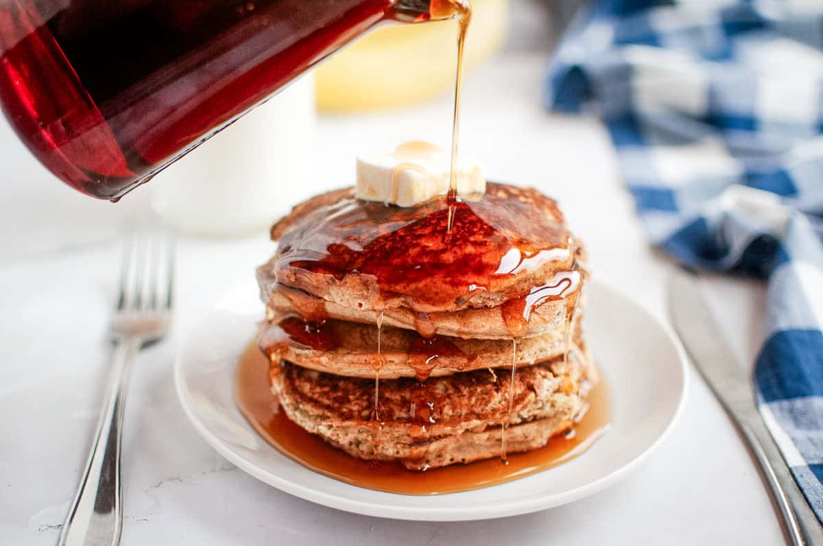 The syrup is poured over a stack of Buttermilk Oatmeal Pancakes topped with butter on a white plate.