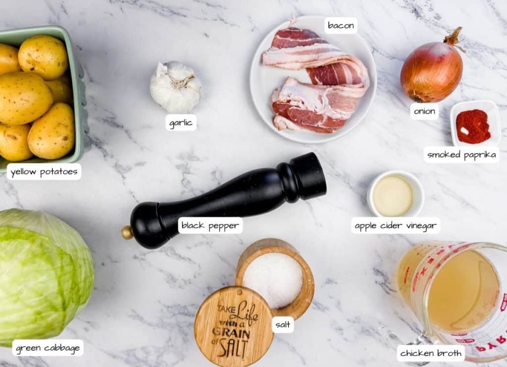 Ingredients for Cabbage & Potatoes with Bacon on a countertop: yellow potatoes, garlic, bacon, onion, smoked paprika, black pepper, apple cider vinegar, salt, chicken broth, and green cabbage.