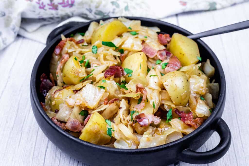 A black bowl filled with Cabbage & Potatoes with Bacon with a spoon on the side.