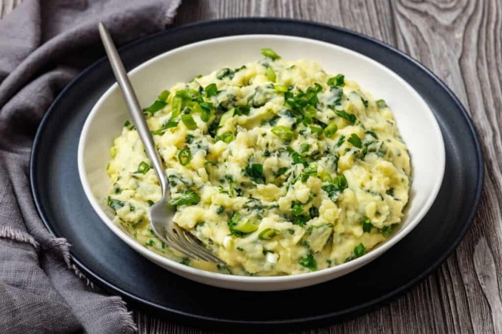 A bowl of Colcannon with Kale with chopped green onions on top, a fork resting inside. The bowl is on a black plate, set on a wooden surface with a gray cloth nearby.