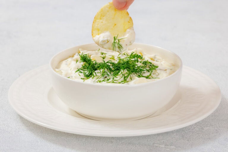 A hand dips a potato chip into a bowl of dill pickle dip topped with fresh dill.