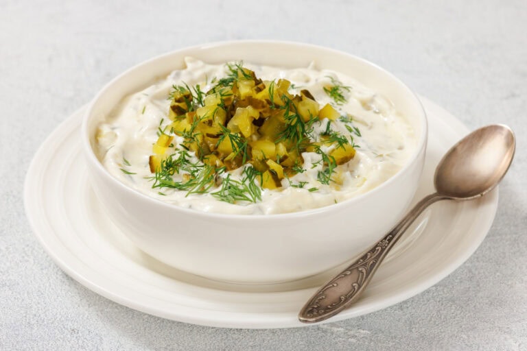A bowl of dill pickle dip topped with diced pickles and fresh dill, placed on a white plate with a spoon beside it.