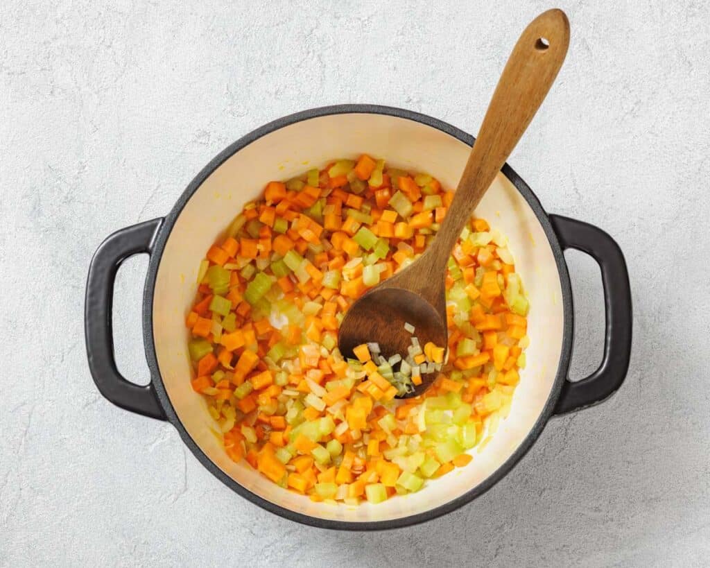 A pot with diced carrots, celery, and onions being stirred with a wooden spoon.