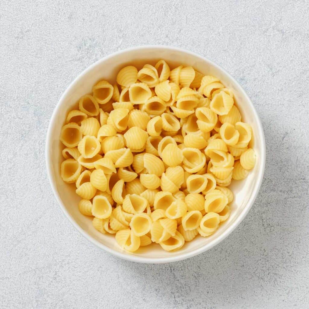 A white bowl filled with uncooked, shell-shaped pasta on a textured light gray background.