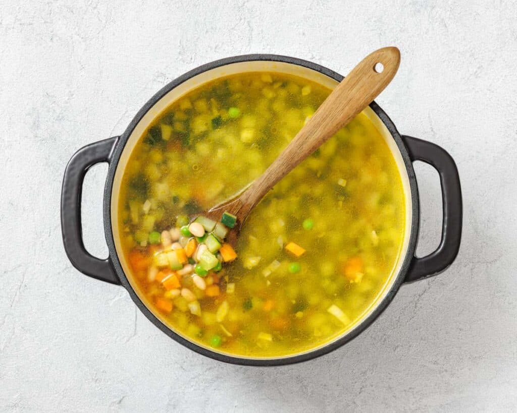 A pot of vegetable soup with a wooden spoon, containing diced carrots, peas, and beans, viewed from above.