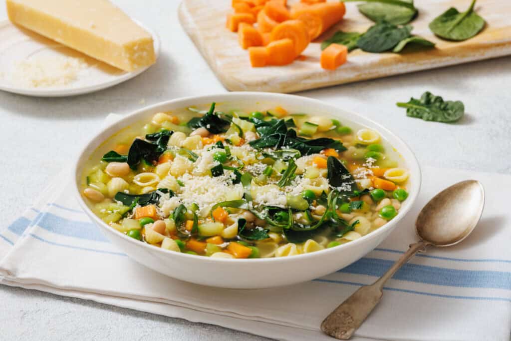 A bowl of Spring Minestrone with pasta, spinach, peas, and beans, garnished with grated cheese. A wooden board with sliced carrots and spinach, and a piece of cheese in the background.