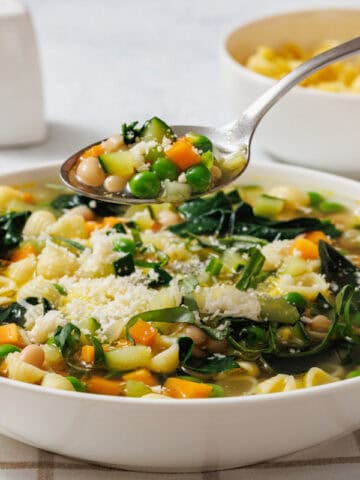 A bowl of Spring Minestrone with pasta, peas, carrots, beans, and greens. A spoon holding some soup is above the bowl. Salt and pepper shakers are in the background.