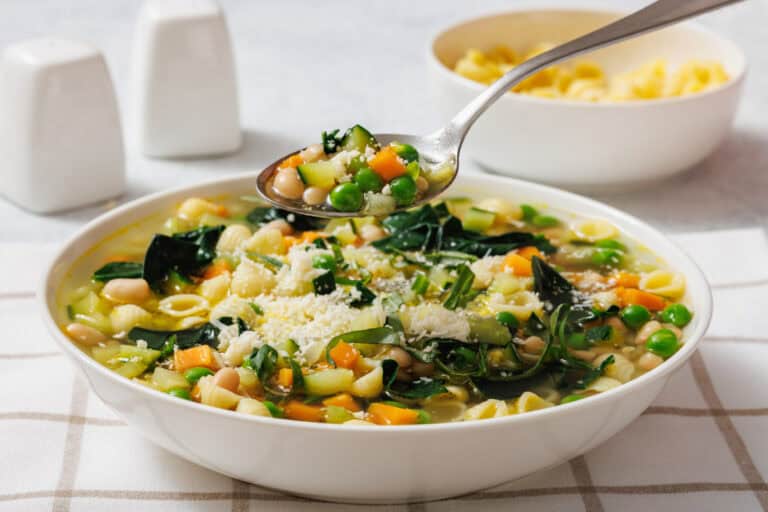A bowl of spring minestrone with pasta, peas, carrots, beans, and greens. A spoon holding some soup is above the bowl.