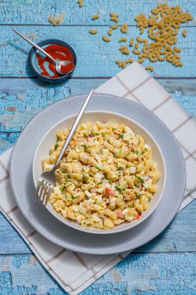 A bowl of Deviled Egg Pasta Salad with diced vegetables and herbs on a white plate, accompanied by a fork. A small dish of red spice and scattered pasta shapes are in the background.