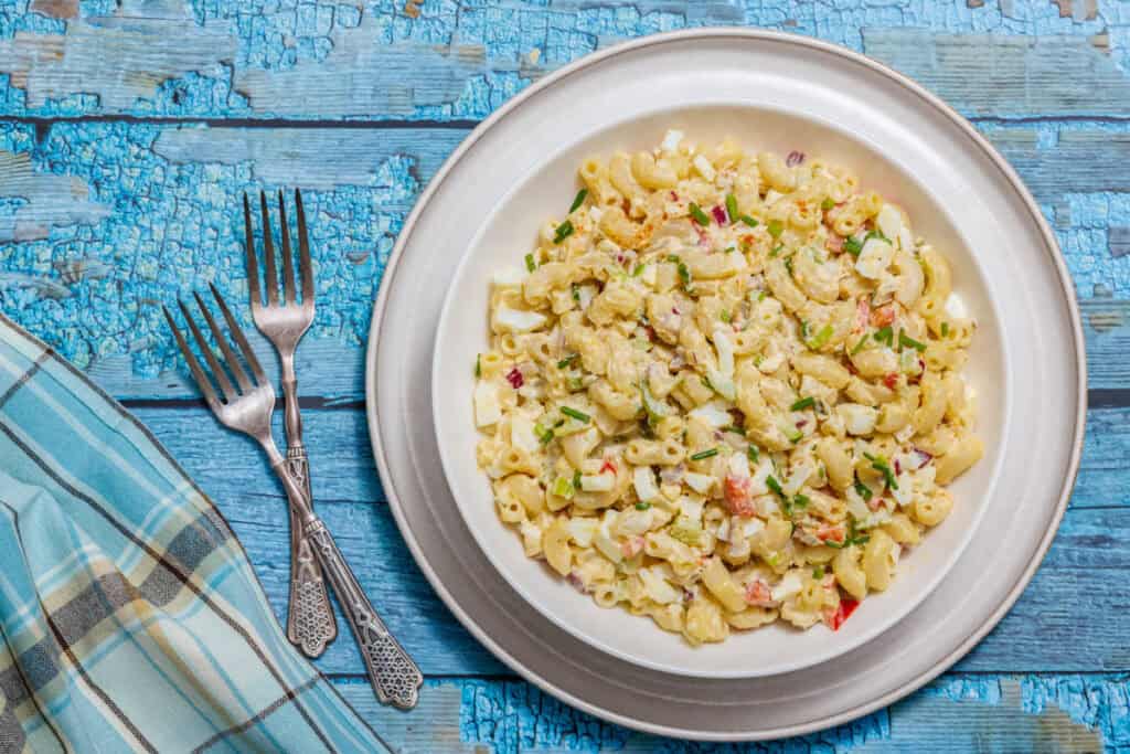 Plate of Deviled Egg Pasta Salad on a wooden table, accompanied by three forks and a blue plaid napkin.