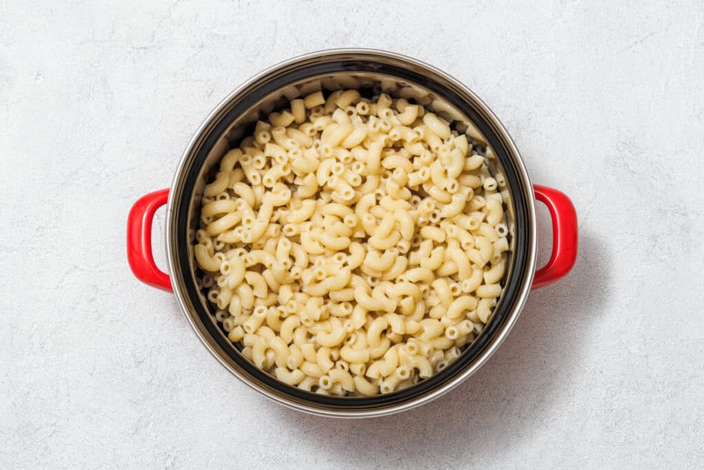 A pot of cooked elbow macaroni with red handles on a light gray surface.
