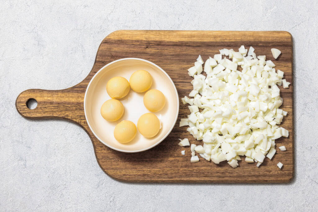 A wooden cutting board with a bowl containing five peeled boiled eggs and a pile of chopped egg whites beside it.