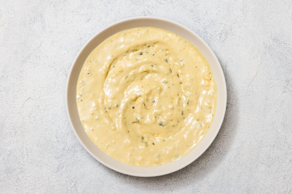 A bowl of creamy mustard sauce with herbs on a textured light gray surface.
