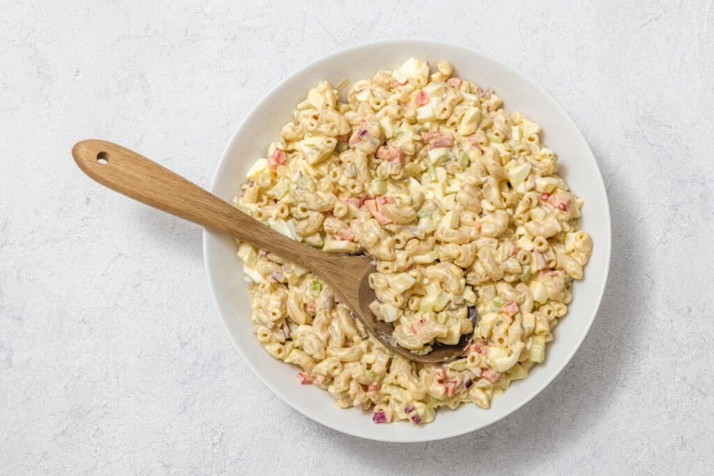A bowl of macaroni salad with a wooden spoon. The salad contains pasta, diced vegetables, and a creamy dressing, placed on a light gray surface.