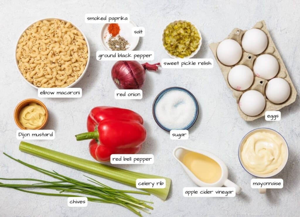Ingredients for a Deviled Egg Pasta Salad on a white surface: elbow macaroni, eggs, red bell pepper, celery rib, chives, Dijon mustard, mayonnaise, smoked paprika, salt, sugar, relish, vinegar, pepper.