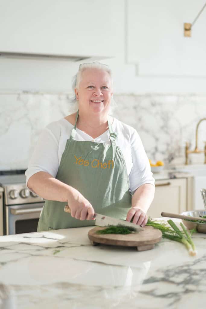 Chef Jenn Allen in a kitchen wearing a green apron with "Yes Chef" text, smiling while chopping herbs on a wooden board. Bright, modern kitchen with marble countertops and brass fixtures in the background.