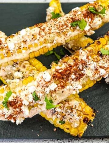 Grilled corn on the cob pieces topped with cheese, chili powder, and cilantro, served on a black slate plate with lime wedges on the side.