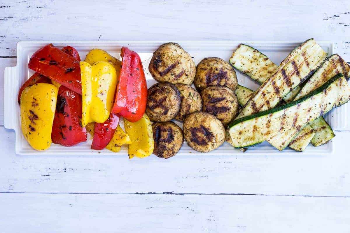 Grilled vegetables on a rectangular white platter, including red and yellow bell peppers, mushrooms, and zucchini slices.