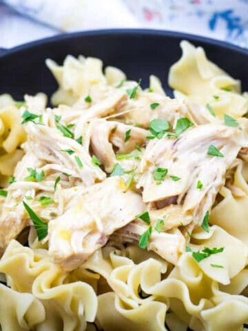 A black bowl containing egg noodles topped with Slow Cooker Mississippi Chicken and garnished with parsley, accompanied by a pickled pepper.