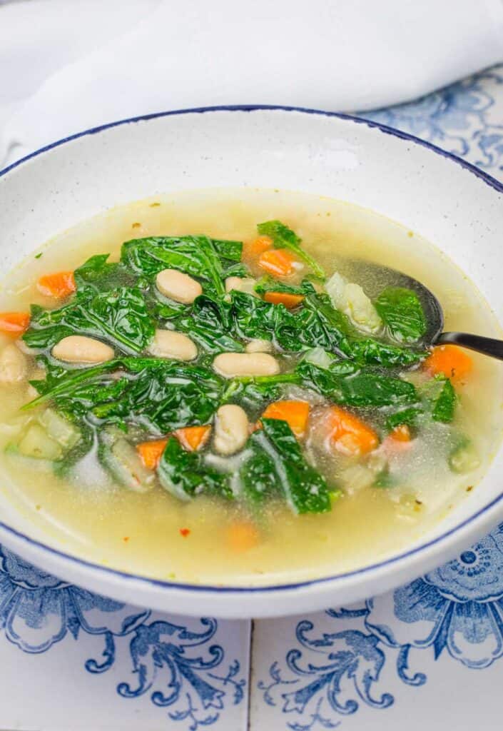 A bowl of Rice and White Bean Soup containing white beans, kale, carrots, and other vegetables, with a spoon on the side.