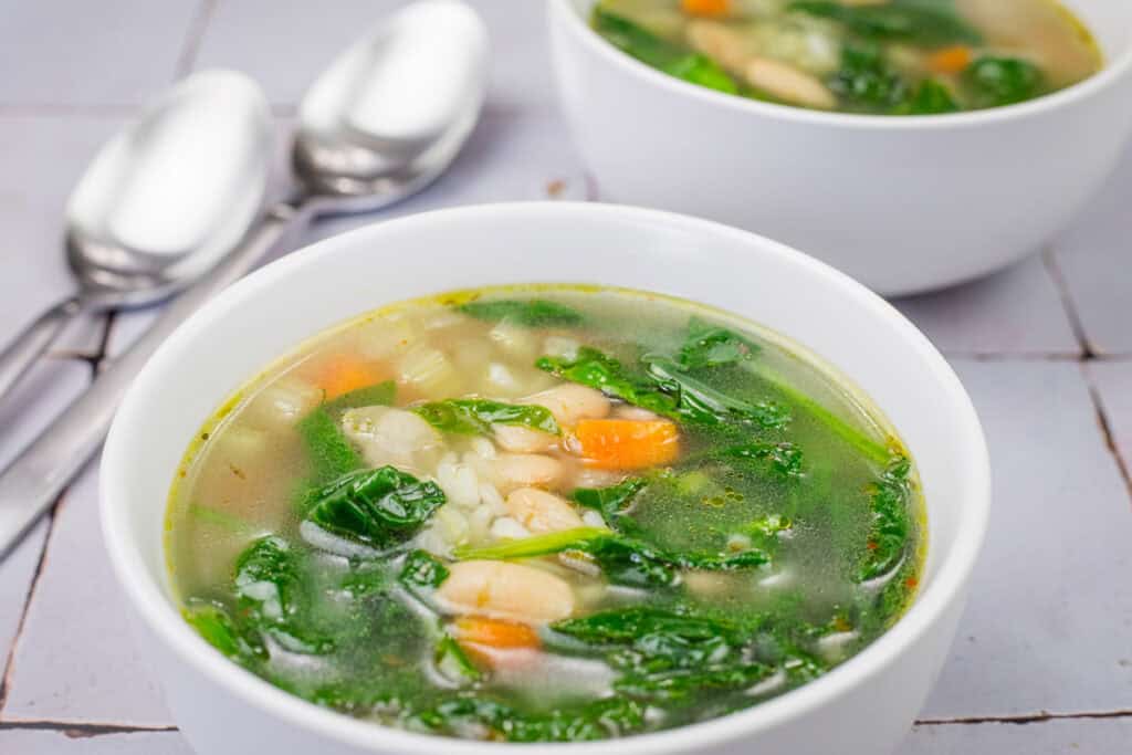 Bowl of Rice and White Bean Soup with spinach, carrots, and beans on a tiled surface, with two spoons nearby.