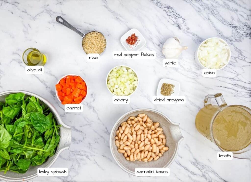 Ingredients for a Rice and White Bean Soup recipe displayed on a marble surface: olive oil, rice, red pepper flakes, garlic, onion, carrot, celery, dried oregano, broth, cannellini beans, baby spinach.
