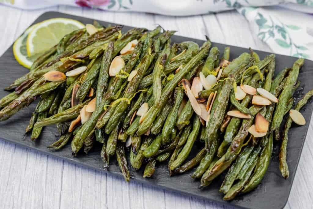 A plate of roasted green beans garnished with sliced almonds, accompanied by lemon slices on a black serving board.