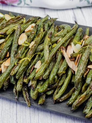 A plate of roasted green beans garnished with sliced almonds, accompanied by lemon slices on a black serving board.