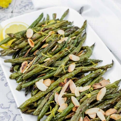 A rectangular plate of roasted green beans garnished with sliced almonds and lemon wedges on a patterned table surface.