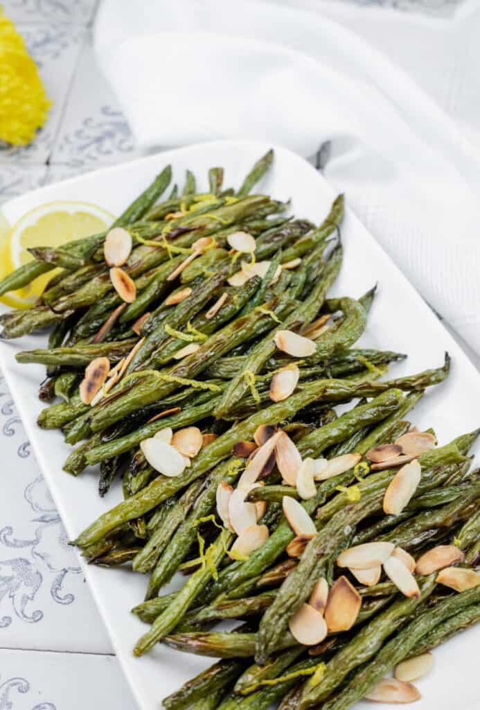 A rectangular plate of roasted green beans garnished with sliced almonds and lemon wedges on a patterned table surface.