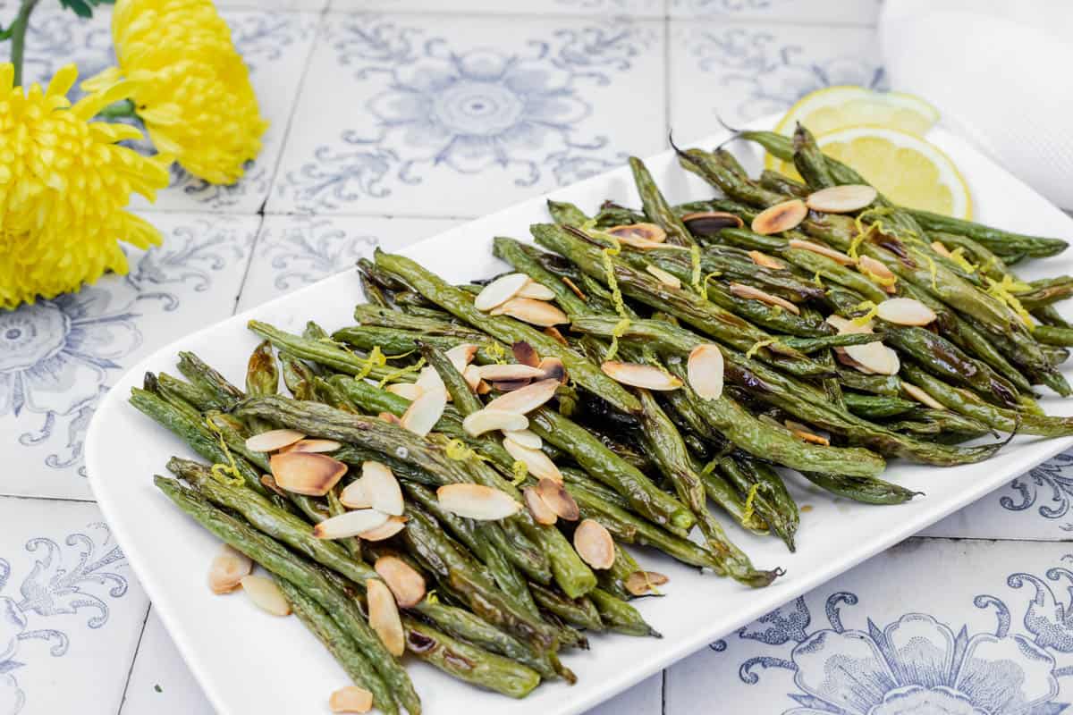 A plate of roasted green beans topped with sliced almonds, garnished with lemon slices on a decorative tiled surface. Yellow flowers are visible in the background.
