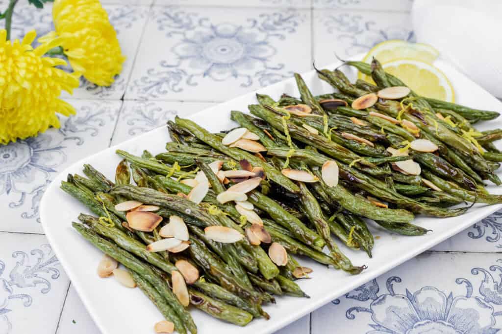 A plate of roasted green beans topped with sliced almonds, set on a patterned surface with yellow flowers and lemon slices in the background.