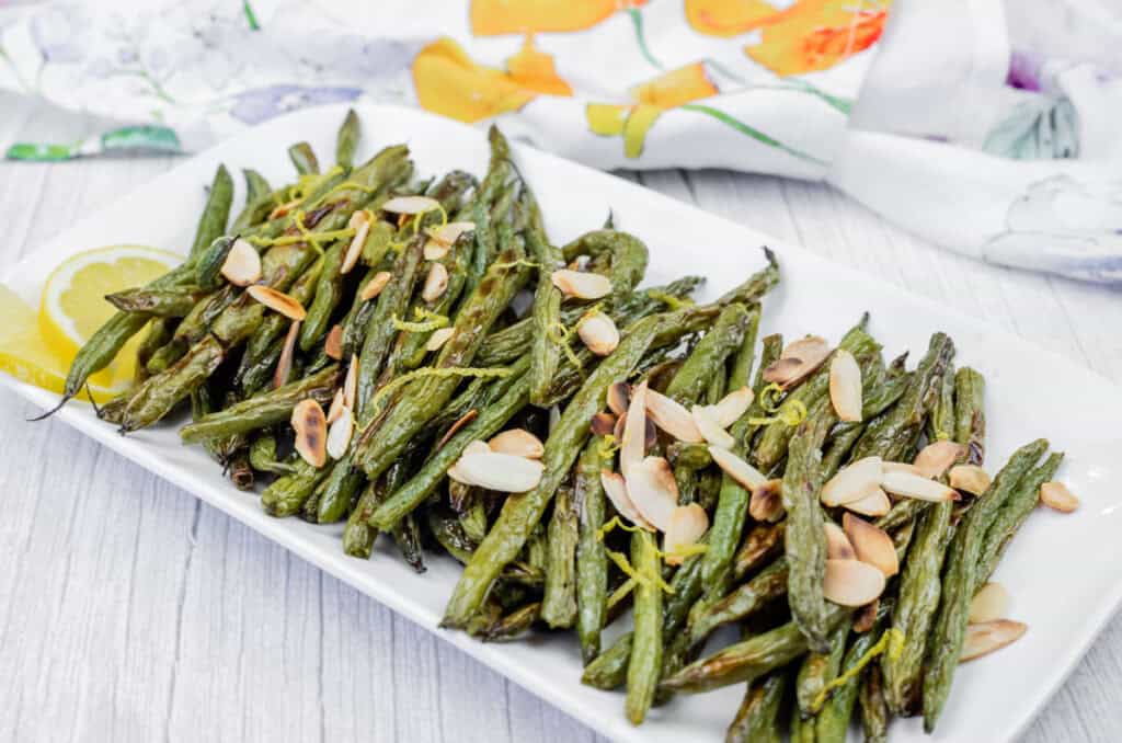 Plate of cooked green beans topped with sliced almonds, garnished with a lemon wedge, on a white rectangular dish. Bright floral cloth in the background.