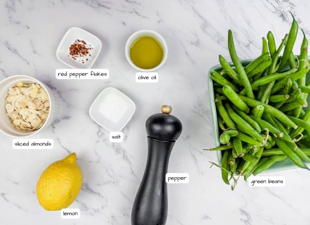 Green beans, olive oil, salt, sliced almonds, red pepper flakes, lemon, and a pepper mill arranged on a marble surface.