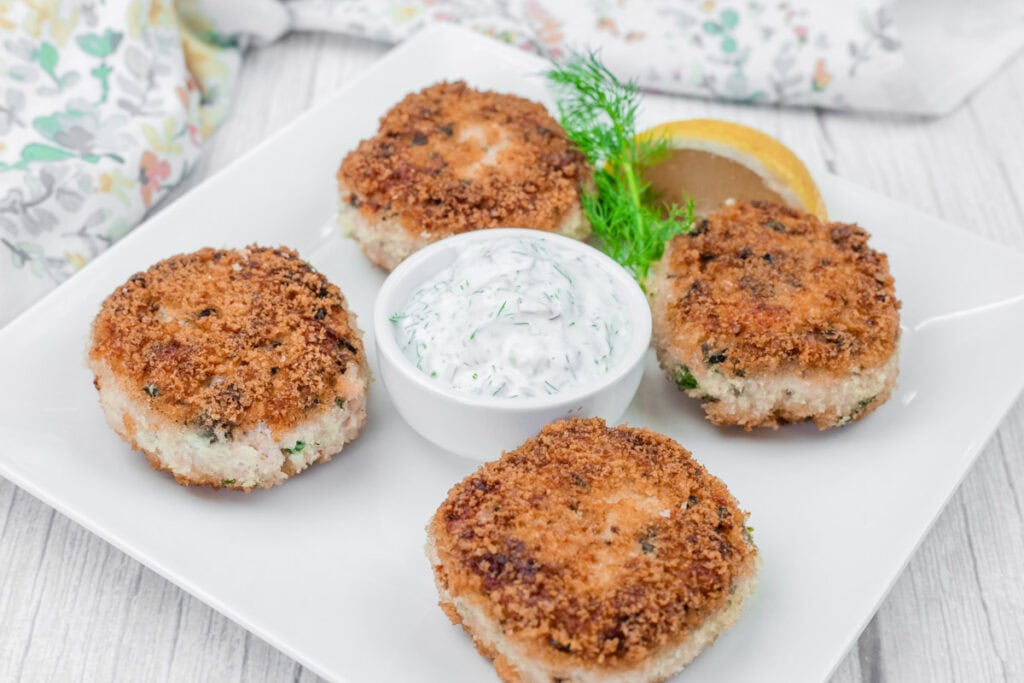 Four Salmon Cakes on a white plate with a small dish of dill sauce, a lemon wedge, and a sprig of dill for garnish.