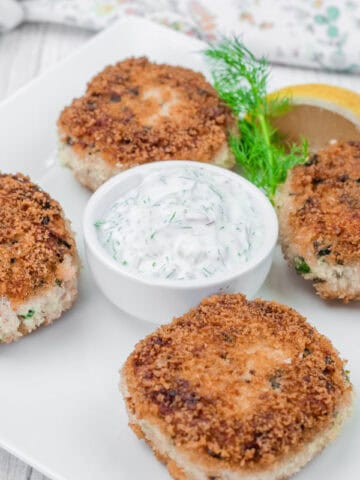 Four Salmon Cakes on a white plate with a small dish of dill sauce, a lemon wedge, and a sprig of dill for garnish.