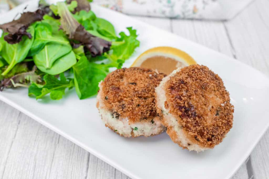 Two Salmon Cakes on a white plate with mixed leafy greens and a lemon wedge.