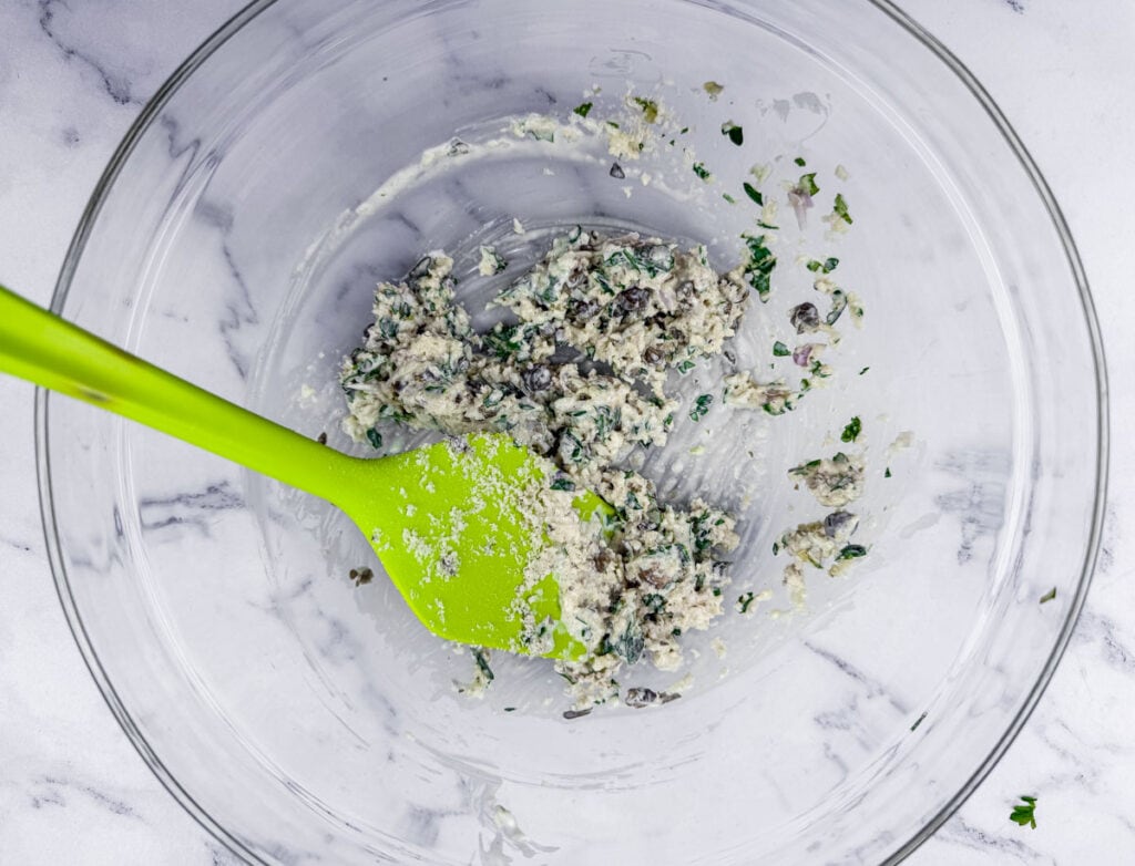 Green spatula in a glass bowl with a mixture of black beans, herbs, and other ingredients on a marble surface.