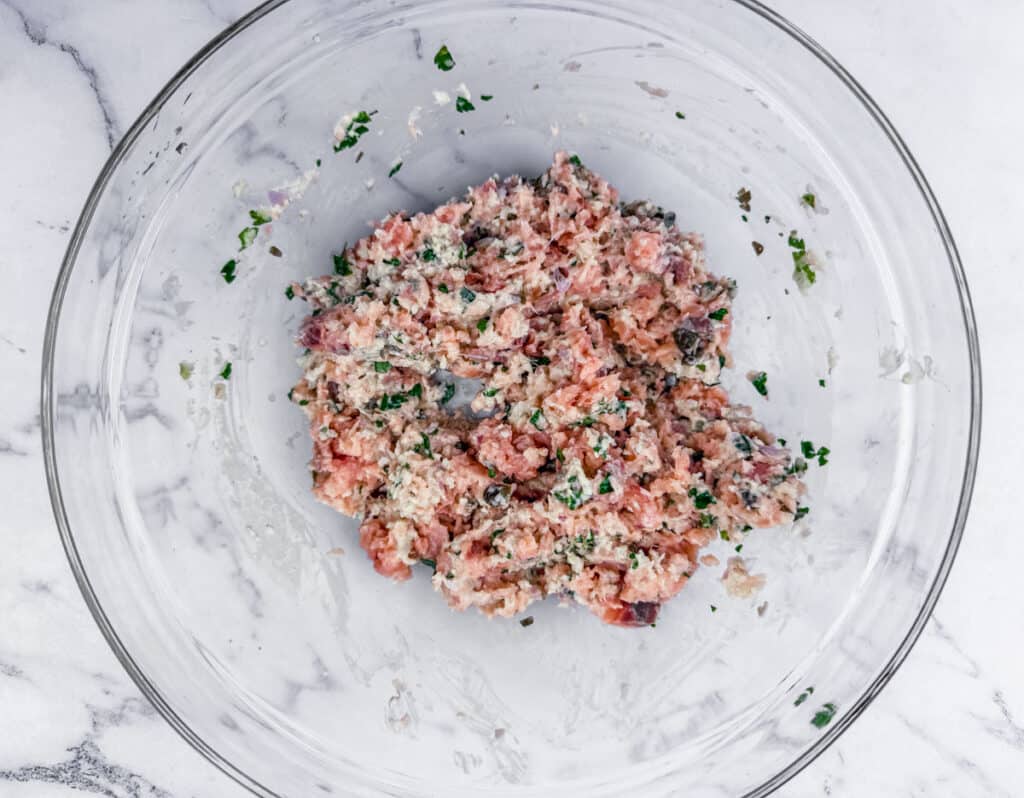A glass bowl contains a mixture of ground meat blended with herbs and spices on a marble countertop.