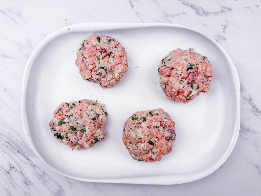 Four raw hamburger patties with herbs on a white oval plate, set atop a marble countertop.