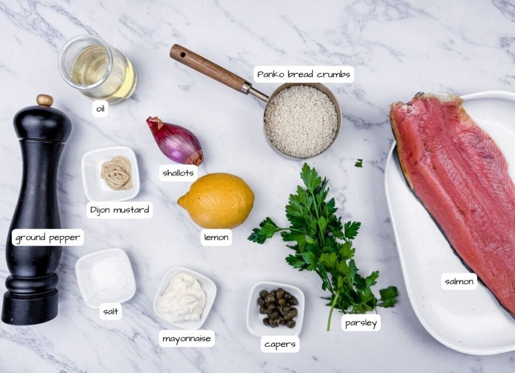 Salmon with ingredients: panko bread crumbs, oil, ground pepper, mustard, shallots, lemon, capers, parsley, mayonnaise, and salt on a white marble surface.