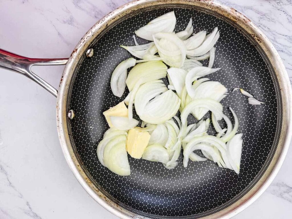 Sliced onions and butter in a non-stick frying pan on a marble countertop.